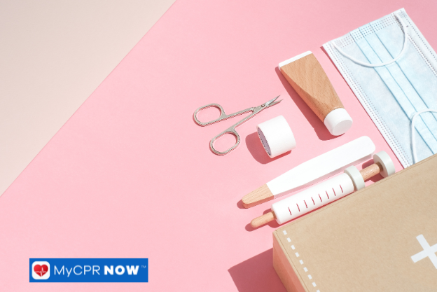 A pastel pink background with wooden and plastic first aid tools, including a mask, scissors, and bandages.