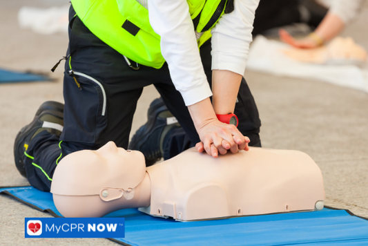 A rescue worker in red safety gear performs CPR on a manikin outdoors, indicating emergency preparedness in a public setting.