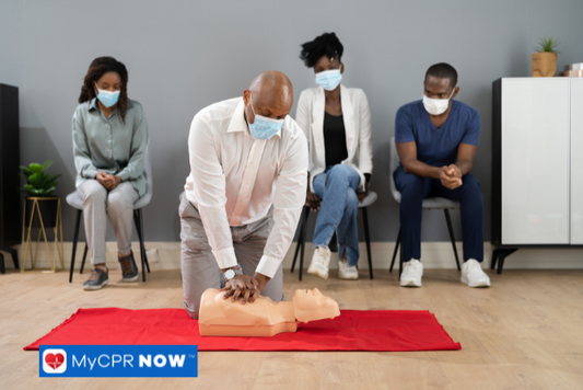 CPR training session where a man in a white shirt performs chest compressions on a manikin while participants wearing masks observe.