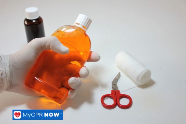 A gloved hand holding an orange antiseptic bottle next to bandages and medical scissors.