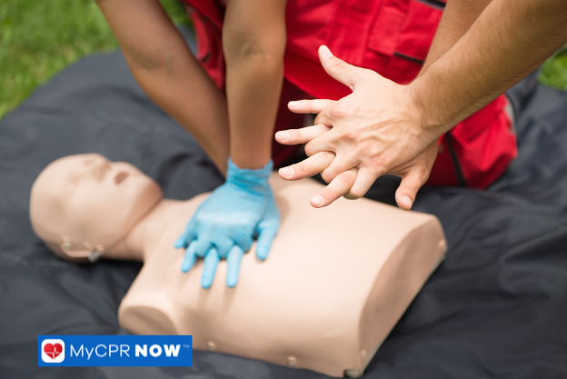 A person in red uniform performing CPR on a mannequin outdoors, with another individual assisting.