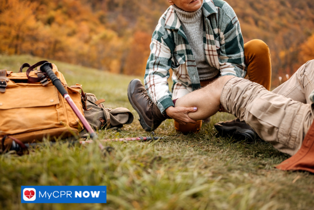 A person helping another with a leg injury on the grass beside hiking gear in an outdoor setting.