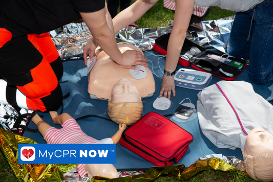 Two people are conducting CPR on a training manikin surrounded by a first aid kit, bandages, and tape