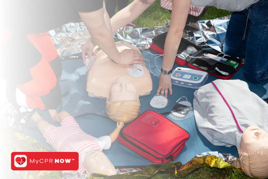 Two people on a blanket in the grass with CPR three mannequins and AEDs.  