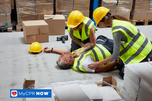 Two workers in safety vests assisting an unconscious colleague in a warehouse.