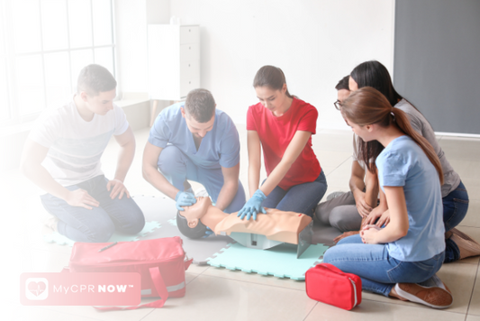 A group of people around an adult CPR mannequin, two people in blue gloves performing cpr on it. 