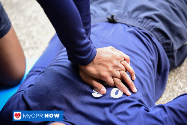 Hands performing CPR compressions on an individual in a blue uniform lying on the ground.