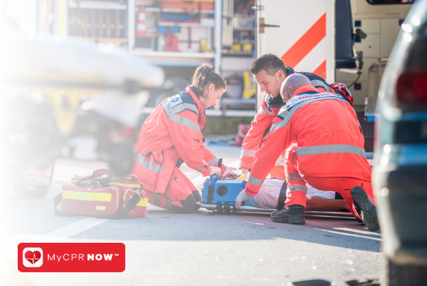 Medical professionals knelt down arounds somone on the ground outside with a stretcher and an open ambulance behind them . 