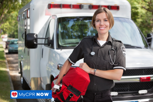 A smiling paramedic standing in front of an ambulance holding a red first aid kit.