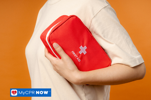 Person holding first aid kit – Someone holding a red first aid kit against their chest with an orange background.