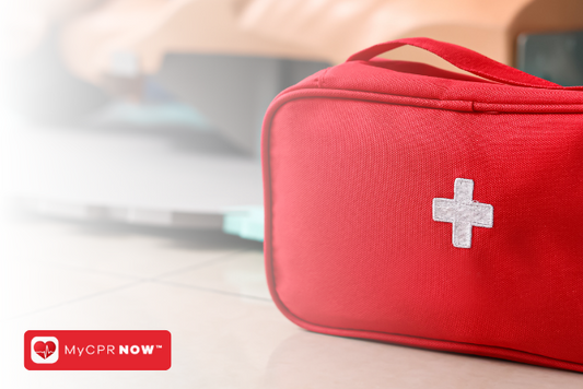 A red bag with a white first aid cross on the front, set in front of a CPR mannequin in the distance. 