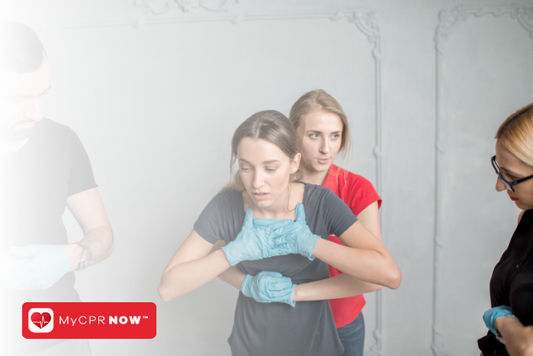 A woman in a red shirt and blue gloves performing the Heimlich Maneuver on another woman wearing a dark blue shirt and blue gloves with her hands to her throat, and two people off to either side. 