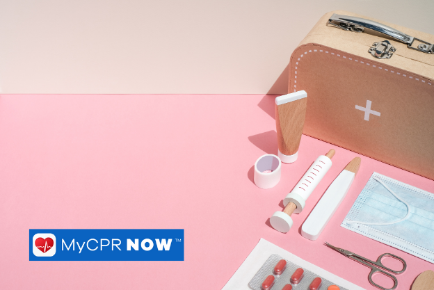 A First Aid kit containing a syringe, scissors, mask, medicine, cream, and bandages is laid on a pink table. 