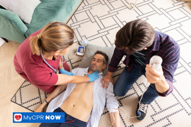 A nurse assisting a person lying on the floor while another individual observes with a defibrillator.