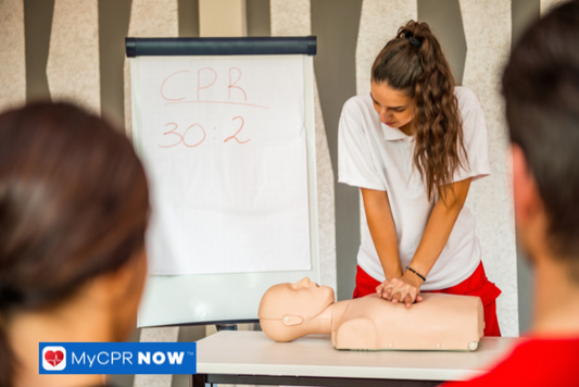 A CPR instructor demonstrating chest compressions on a manikin with a whiteboard in the background.