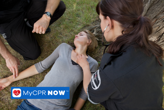Two first responders kneeling next to a woman laying in the grass next to a tree checking her pulse. 