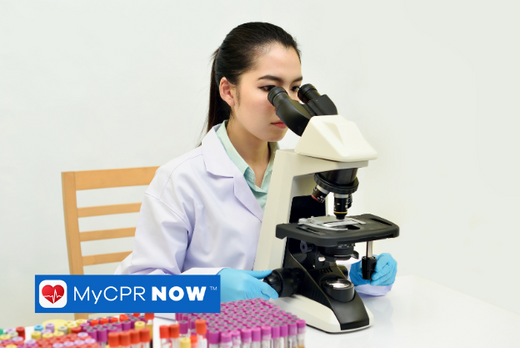 A medical professional loocking throuhg a microscope with viles on the table next to the microscope.