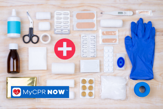 First aid supplies like gloves, medical tape, a thermometer, adhesive bandages, scissors, and ointments on a wooden surface. 