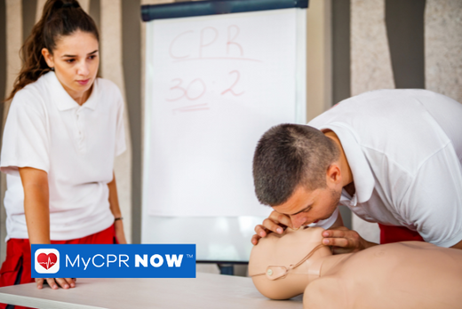 A man in a classroom performing mouth to mouth on a adult CPR mannequin that is on a table, while a woman stands to his right watching and a whiteboard behind her that has written on it "CPR 30 2".  