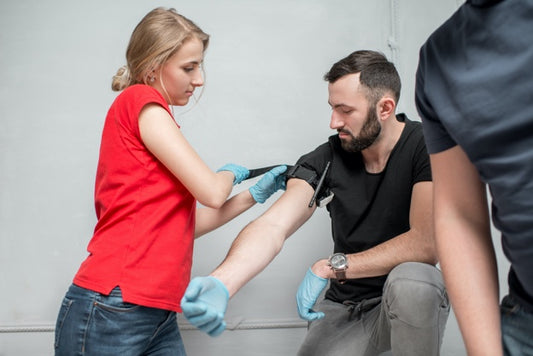 A woman in a red shirt and blue gloves applying a tourniquet on a mans right arm with another person half off the frame. 